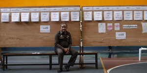 Ein Polizist sitzt auf einer Bank. Hinter ihm Wählerlisten
