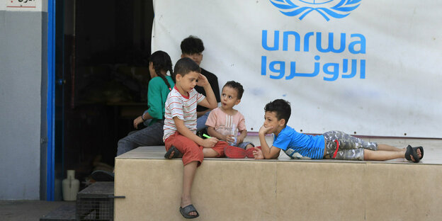 Kinder sitzen und liegen vor einem Plakat mit Aufschrift UNRWA