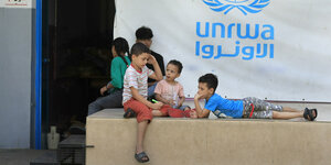 Kinder sitzen auf einem Betonsockel, vor einer Plane mit dem Logo der URNWA